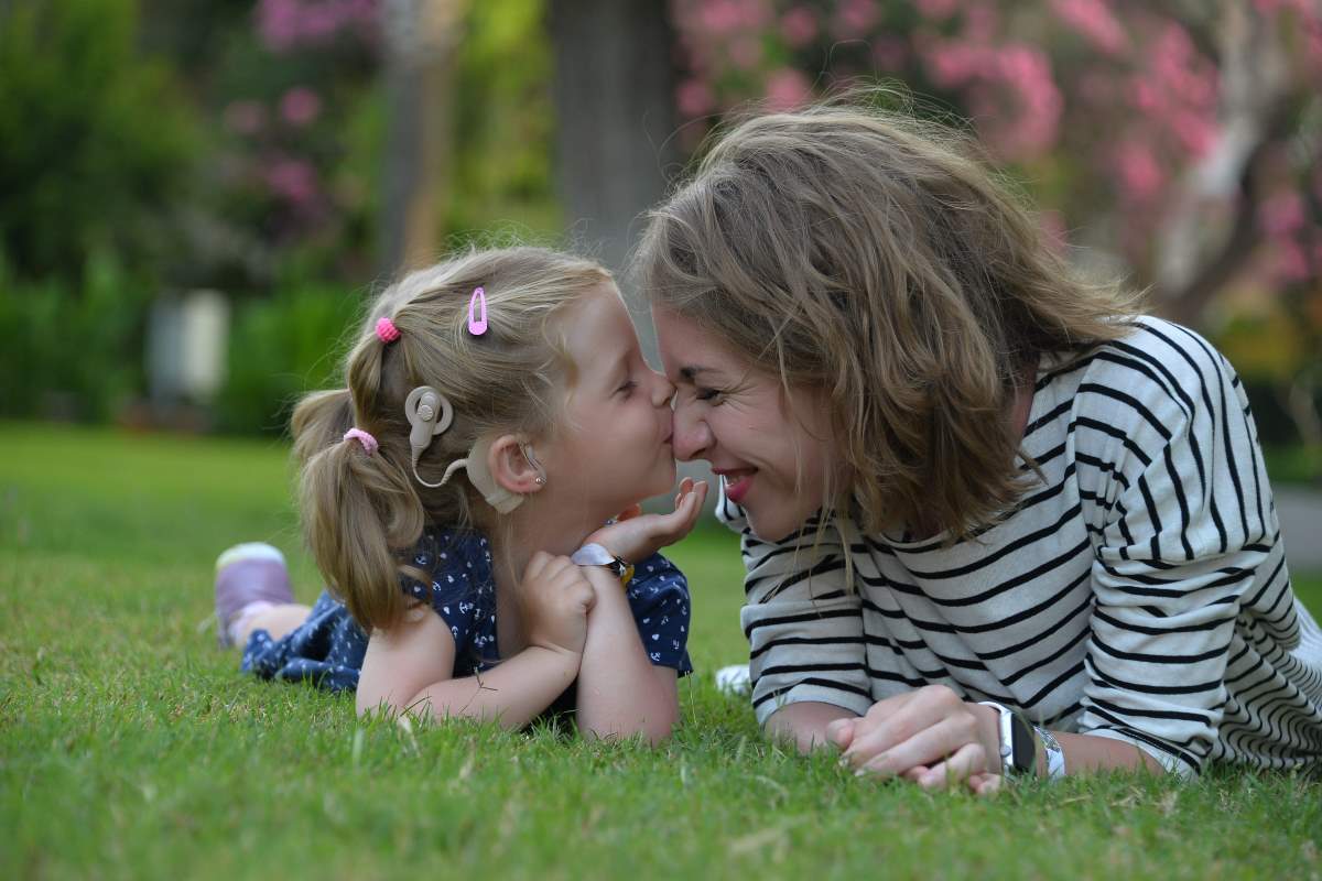 Девочка с кохлеарным имплантом целует свою маму , лежа на лужайке